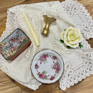 Pu&ograve; includere: Un assortimento di oggetti vintage su una tovaglia di pizzo. Include una piccola scatola decorativa con una scena storica, un timbro per sigillo di cera color oro, una candela bianca, un piccolo piatto floreale e una rosa bianca.