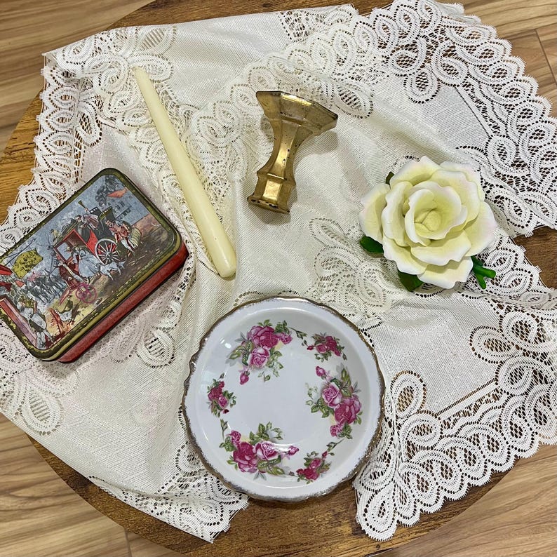 Pu&ograve; includere: Un assortimento di oggetti vintage su una tovaglia di pizzo. Include una piccola scatola decorativa con una scena storica, un timbro per sigillo di cera color oro, una candela bianca, un piccolo piatto floreale e una rosa bianca.