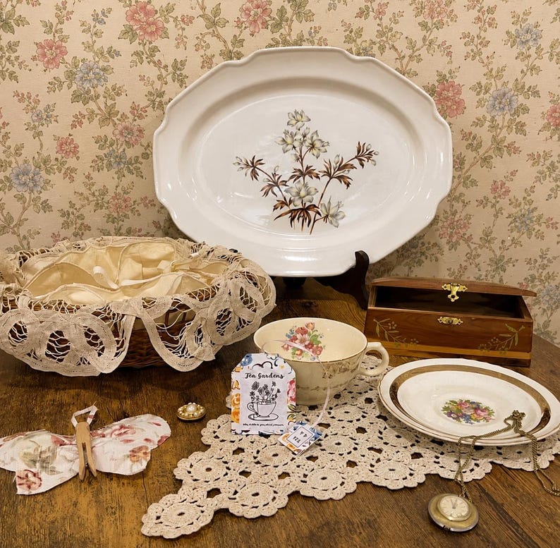 Pu&ograve; includere: Una collezione di oggetti vintage tra cui un grande piatto ovale bianco con un motivo floreale, un cestino intrecciato con finiture in pizzo, una tazza da t&egrave;, una piccola scatola di legno e un orologio da tasca. Sono presenti anche una decorazione a farfalla e un centrino.