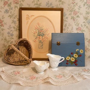 Pu&ograve; includere: Una natura morta con un cestino di vimini a forma di cuore, una stampa floreale incorniciata, una scatola di legno blu con fiori dipinti e due lattine di vetro bianco latte. La scena &egrave; ambientata su una tovaglia a motivi floreali.