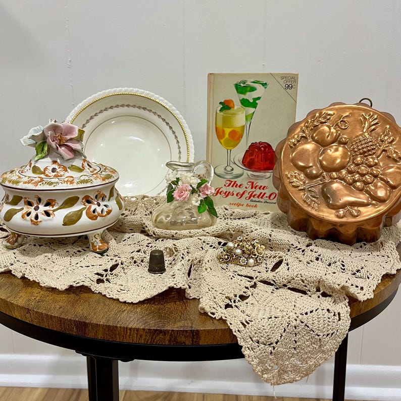 Pu&ograve; includere: Una natura morta su un tavolo rotondo presenta oggetti vintage. Sono visibili un piatto in ceramica decorativo con accenti floreali, un piatto bianco con finiture dorate, uno stampo in rame, un libro di cucina vintage e un centrino in pizzo all'uncinetto.
