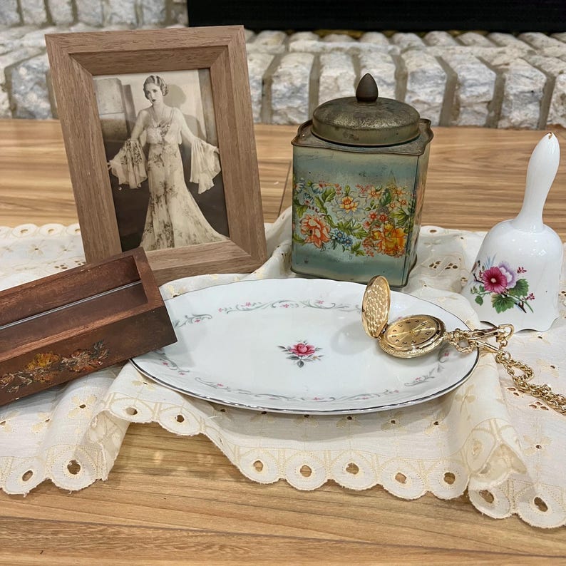 Pu&ograve; includere: Una natura morta vintage con una foto incorniciata, una scatola di t&egrave; decorata con fiori, una campana bianca e un orologio da tasca dorato. Sono presenti anche un piatto ovale bianco con una rosa rosa e una scatola di legno.