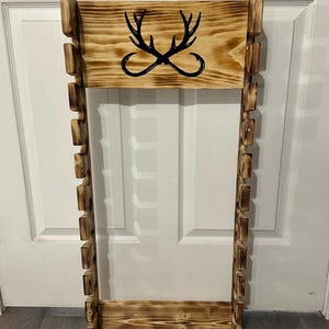 May include: Wooden wall-mounted rack with a burnt finish. The rack features a rectangular top with a black antler and hook design. The sides have multiple rectangular cutouts. A rustic, decorative piece.