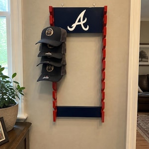 May include: A wall-mounted wooden hat rack featuring a dark blue top panel with a white letter A logo. The frame is red with multiple pegs on both sides. Four dark gray baseball caps are hanging on the left side of the rack in a vertical stack.