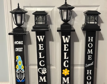 May include: Four black vertical wooden porch signs with solar-powered lanterns on top. Each sign features white lettering, including phrases like HOME SWEET GNOME, WELCOME with a paw print, WELCOME with a yellow flower, and HOME SWEET HOME.