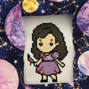 May include: Cross-stitch art of a cartoon character in a purple dress holding a bloody knife. The cross-stitch is framed by fabric with a dark blue background and celestial patterns, including planets and stars.