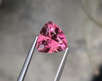 May include: A faceted, triangular pink gemstone is held by silver tweezers. The gem has a vibrant, saturated color and a polished surface, reflecting light. The background is a soft, blurred neutral tone.