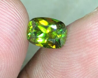 May include: A green gemstone with a faceted cut. The gemstone is oval shaped and has a golden hue.