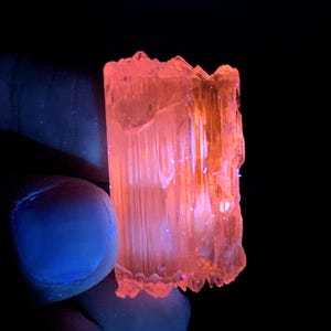 Peut inclure: Un cristal translucide orange-rose avec une texture stri&eacute;e. Le cristal est &eacute;clair&eacute; par une lumi&egrave;re UV, ce qui le fait briller intens&eacute;ment.
