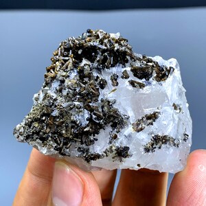 May include: A close-up of a mineral specimen featuring a clear, crystalline base with clusters of dark, needle-like crystals. The mineral is held in a hand, showcasing its natural texture and color variations.