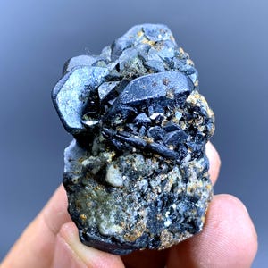 May include: A dark, rough mineral specimen with a mix of black, gray, and gold tones. The surface features multiple crystalline structures and a textured appearance. The mineral is held in a hand against a blurred background.