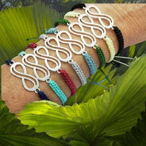Puede incluir: Una colección de pulseras plateadas con el símbolo del infinito y coloridas bandas tejidas. Las pulseras presentan una variedad de colores, incluyendo azul, rojo, verde y negro. Las pulseras se exhiben sobre un fondo de follaje verde.