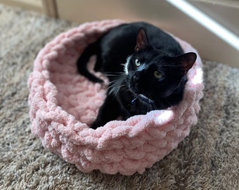 May include: A black cat with a white patch on its chest is resting in a pink knitted pet bed. The bed is round and has a fluffy texture.