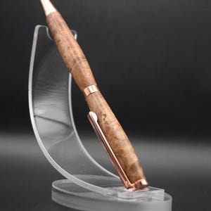 May include: A handcrafted wooden pen with a copper-colored clip and accents. The pen features a light brown, burl wood pattern and a pointed tip. It is displayed on a clear acrylic stand against a dark background.