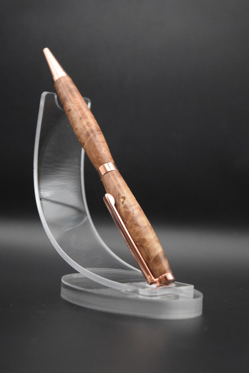May include: A handcrafted wooden pen with a copper-colored clip and accents. The pen features a light brown, burl wood pattern and a pointed tip. It is displayed on a clear acrylic stand against a dark background.