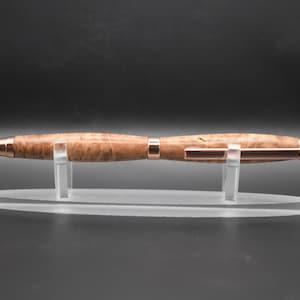 Könnte beinhalten: Ein handgefertigter Holzstift mit hellbraunem, gemustertem Finish und kupferfarbenen Akzenten. Der Stift wird auf einem transparenten Acrylständer präsentiert, der sein elegantes Design hervorhebt. Ein anspruchsvolles Schreibgerät.