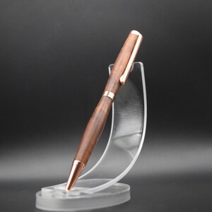 May include: A wooden pen with a rose gold finish and a rose gold clip. The pen is displayed on a clear plastic stand.