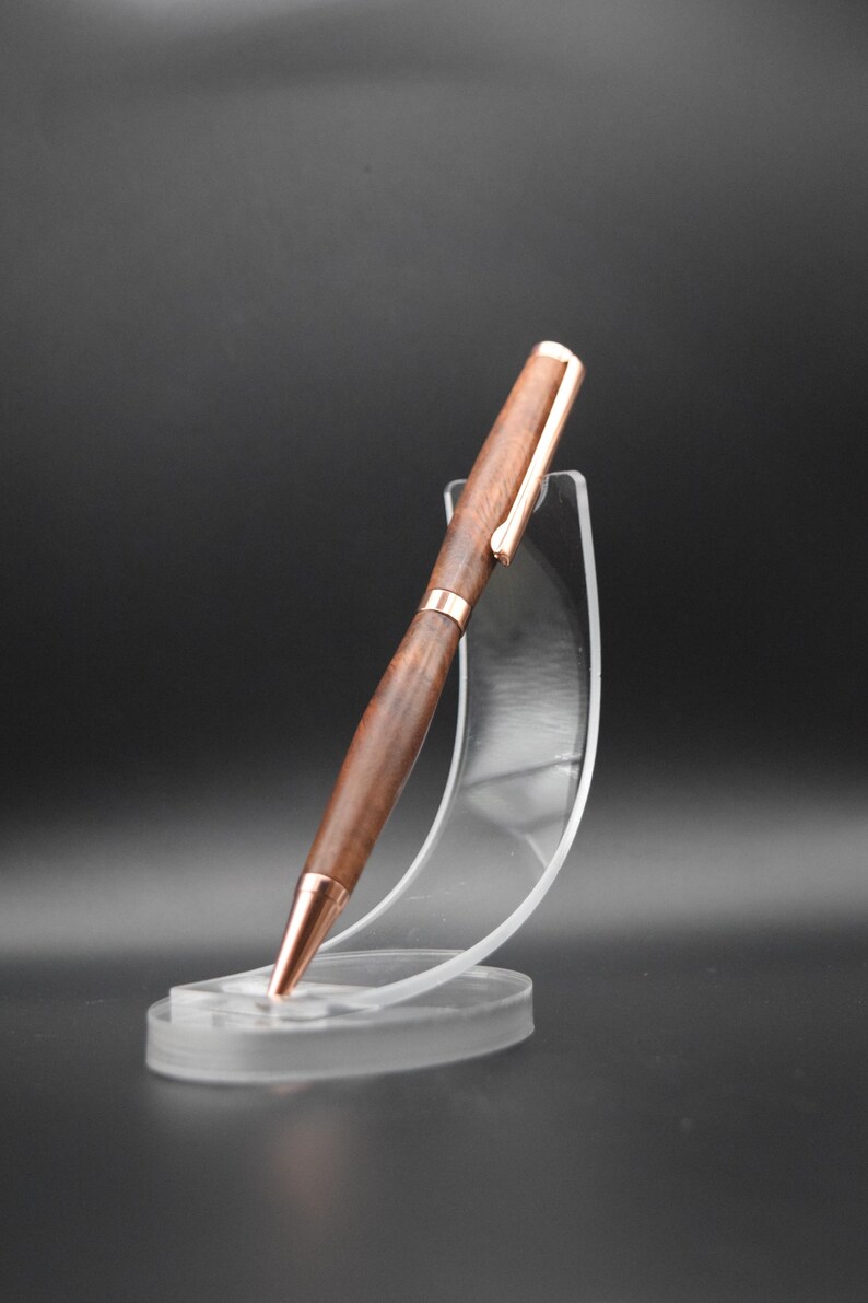 May include: A wooden pen with a rose gold finish and a rose gold clip. The pen is displayed on a clear plastic stand.