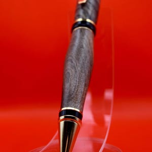 May include: A handcrafted wooden pen with a gray and brown grain pattern. The pen features gold-colored accents on the tip, clip, and bands. The pen is displayed on a clear acrylic stand against a red background.