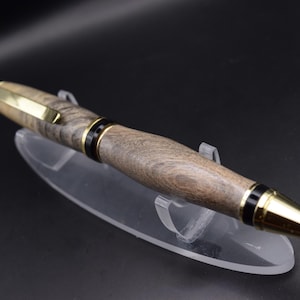 Könnte beinhalten: Ein handgefertigter Holzstift mit goldfarbenem Clip und Spitze. Der Stift hat ein dunkles Holzmaserungsmuster und schwarze Akzente. Der Stift wird auf einem transparenten Acrylständer vor dunklem Hintergrund präsentiert.