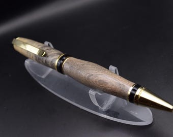 May include: A handcrafted wooden pen with a gold-colored clip and tip. The pen features a dark wood grain pattern and black accents. The pen is displayed on a clear acrylic stand against a dark background.