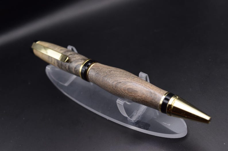 May include: A handcrafted wooden pen with a gold-colored clip and tip. The pen features a dark wood grain pattern and black accents. The pen is displayed on a clear acrylic stand against a dark background.