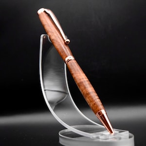 Könnte beinhalten: Ein handgefertigter Holzstift mit kupferfarbenem Clip und Akzenten. Der Stift hat eine dunkelbraune Holzmaserung und wird auf einem transparenten Acrylständer präsentiert. Spitze und Ende des Stifts sind ebenfalls kupferfarben.