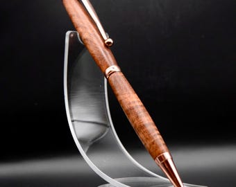 May include: A handcrafted wooden pen with a copper-colored clip and accents. The pen features a rich, brown wood grain pattern and is displayed on a clear acrylic stand. The pen's tip and end are also copper-colored.