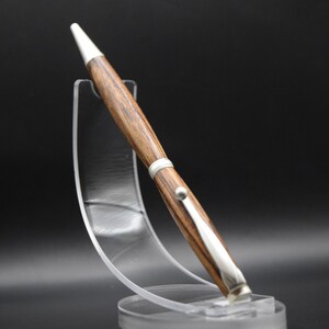 May include: A handcrafted wooden pen with a silver-colored clip and accents. The pen has a white tip and is displayed on a clear acrylic stand against a black background. The wood grain is visible, showcasing a natural pattern.