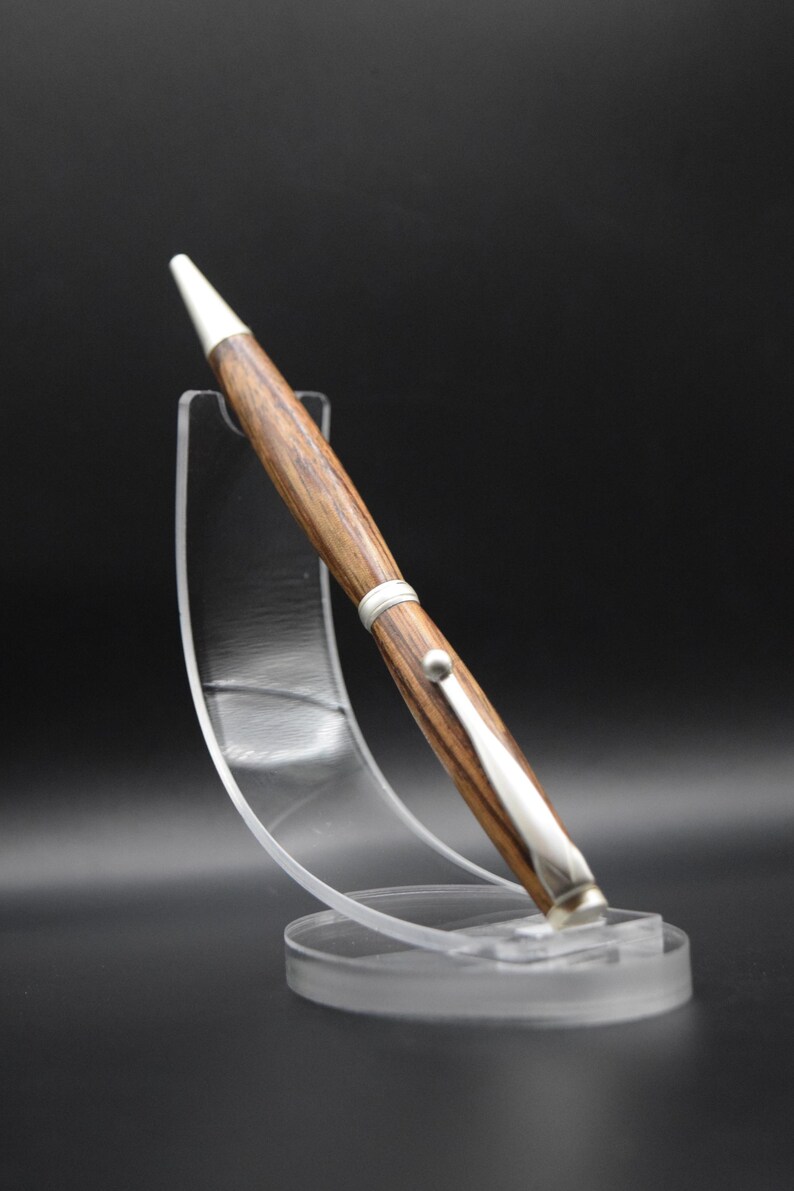 May include: A handcrafted wooden pen with a silver-colored clip and accents. The pen has a white tip and is displayed on a clear acrylic stand against a black background. The wood grain is visible, showcasing a natural pattern.