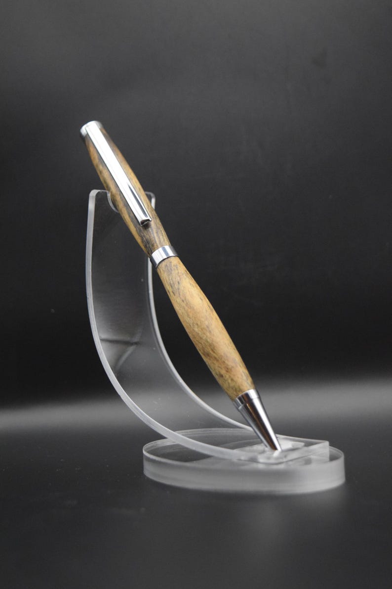 May include: A wooden pen with a silver-colored tip and accents is displayed on a clear acrylic stand. The pen's body has a light brown wood grain pattern. The pen is angled slightly, showcasing its design.