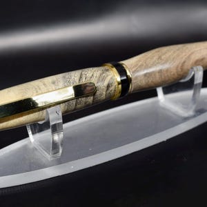 May include: A handcrafted wooden pen with a light brown and gray marbled pattern. The pen features gold-colored accents on the tip, clip, and bands. It rests on a clear acrylic display stand, showcasing its elegant design.
