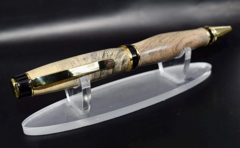 May include: A handcrafted wooden pen with a light brown and gray marbled pattern. The pen features gold-colored accents on the tip, clip, and bands. It rests on a clear acrylic display stand, showcasing its elegant design.
