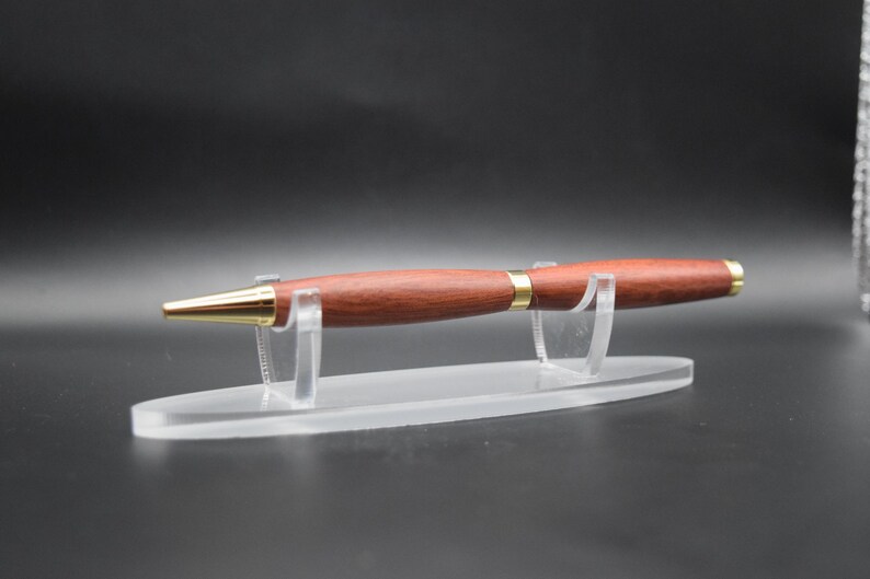 Puede incluir: Un bol&iacute;grafo de madera marr&oacute;n rojizo con detalles dorados, expuesto sobre un soporte de acr&iacute;lico transparente. El bol&iacute;grafo tiene una punta dorada y una banda dorada cerca del centro. Instrumento de escritura.