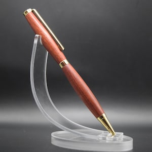 Könnte beinhalten: Ein Holzstift mit rotbraunem Finish und goldfarbenen Akzenten. Der Stift wird auf einem transparenten Acrylständer vor dunklem Hintergrund präsentiert. Das Design des Stifts umfasst einen goldenen Clip und eine goldene Spitze.