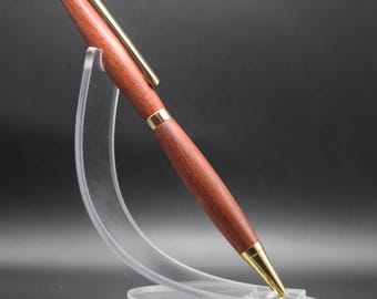 May include: A wooden pen with a reddish-brown finish and gold-colored accents. The pen is displayed on a clear acrylic stand against a dark background. The pen's design includes a gold clip and a gold tip.