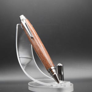 Puede incluir: Un bol&iacute;grafo de madera con un clip y una punta de metal plateado, apoyado en un soporte de pl&aacute;stico transparente.