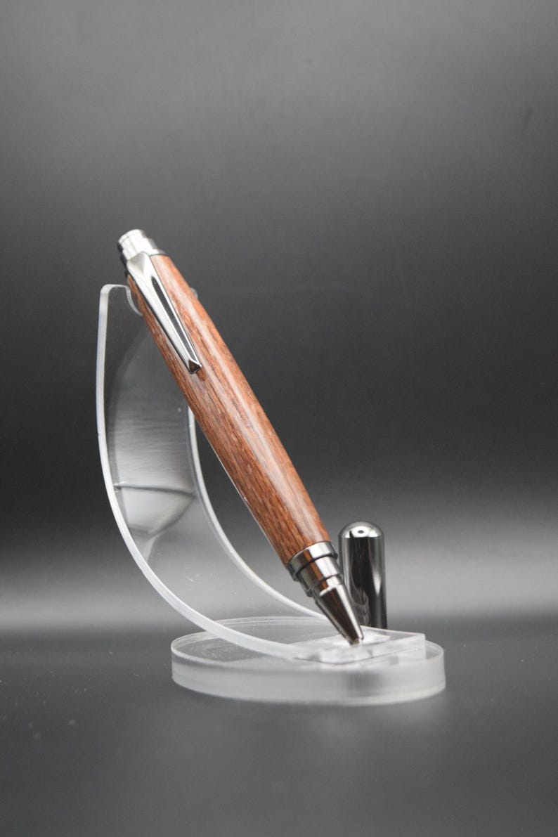 Puede incluir: Un bol&iacute;grafo de madera con un clip y una punta de metal plateado, apoyado en un soporte de pl&aacute;stico transparente.