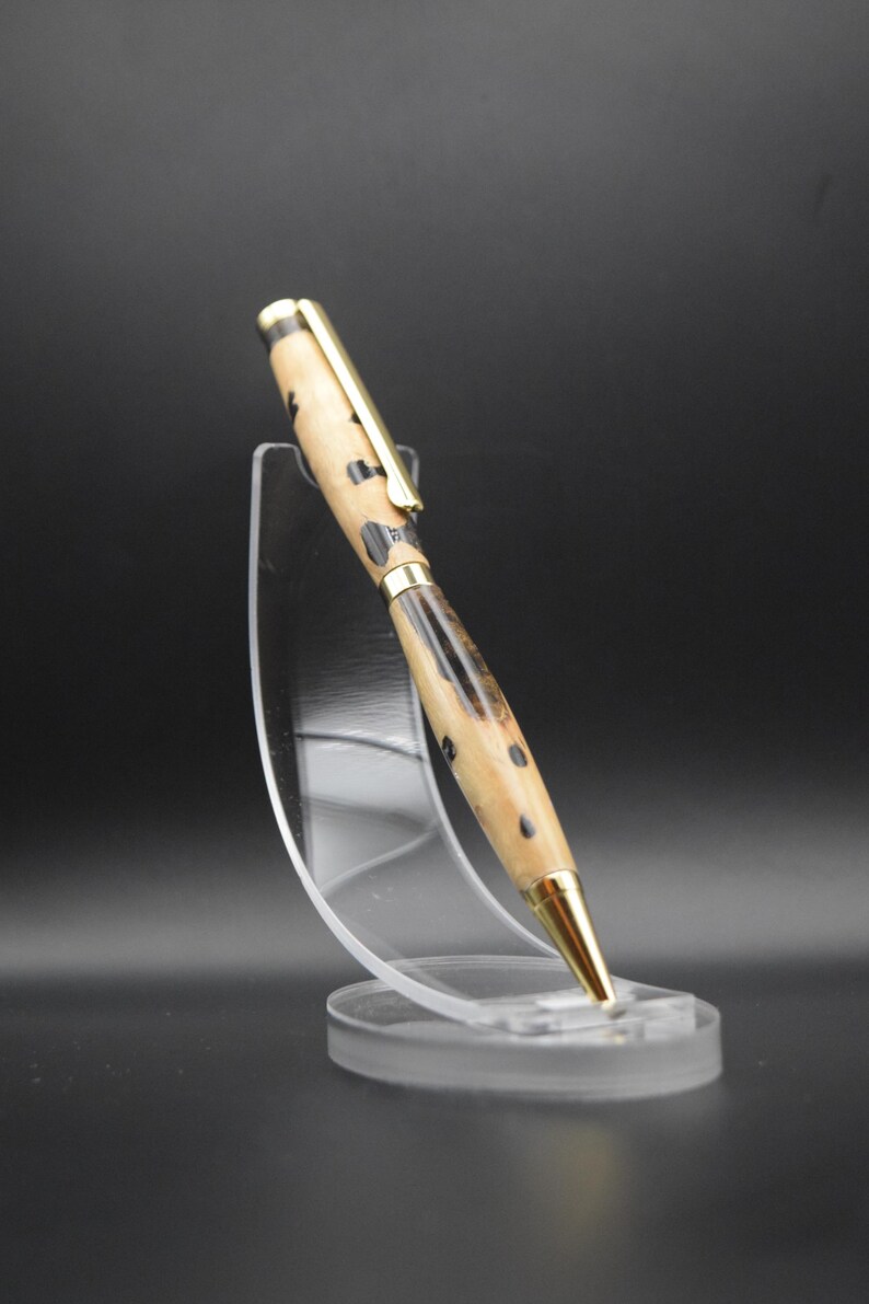 May include: A wooden pen with a gold-colored top and a black and brown wood grain pattern. The pen is displayed on a clear plastic stand.