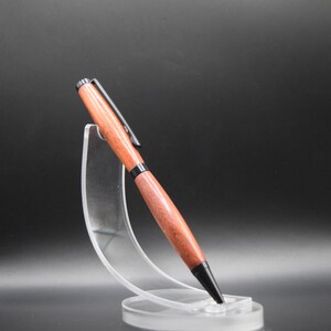 K&ouml;nnte beinhalten: Ein Holzstift mit rotbraunem Finish und schwarzen Akzenten, einschlie&szlig;lich Spitze und Clip. Der Stift wird auf einem transparenten Acrylst&auml;nder vor dunklem Hintergrund pr&auml;sentiert. Das Design des Stifts ist elegant und modern.