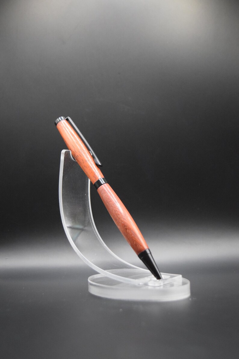 K&ouml;nnte beinhalten: Ein Holzstift mit rotbraunem Finish und schwarzen Akzenten, einschlie&szlig;lich Spitze und Clip. Der Stift wird auf einem transparenten Acrylst&auml;nder vor dunklem Hintergrund pr&auml;sentiert. Das Design des Stifts ist elegant und modern.