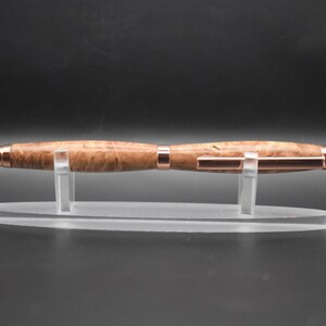 May include: A wooden pen with a light brown finish and copper-colored accents. The pen is displayed on a clear acrylic stand against a dark background. The pen's design features a smooth, polished barrel and a metallic tip.