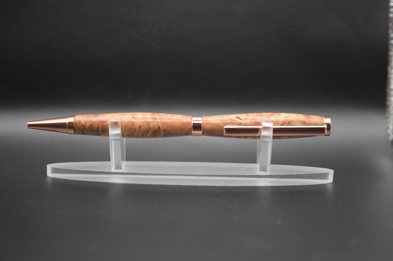 May include: A wooden pen with a light brown finish and copper-colored accents. The pen is displayed on a clear acrylic stand against a dark background. The pen's design features a smooth, polished barrel and a metallic tip.