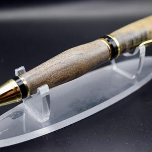May include: A handcrafted wooden pen with a gold-colored tip and accents. The pen features a dark wood grain pattern and is displayed on a clear acrylic stand. The pen's design includes black and gold details, creating a sophisticated writing instrument.