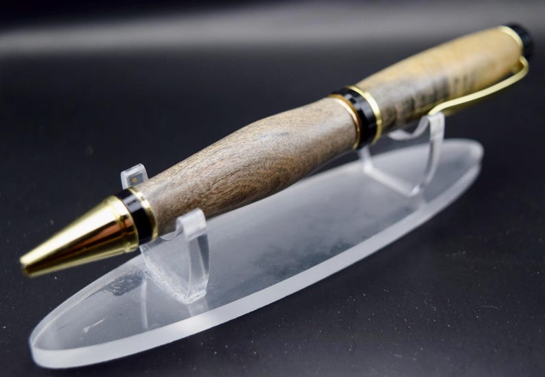 May include: A handcrafted wooden pen with a gold-colored tip and accents. The pen features a dark wood grain pattern and is displayed on a clear acrylic stand. The pen's design includes black and gold details, creating a sophisticated writing instrument.