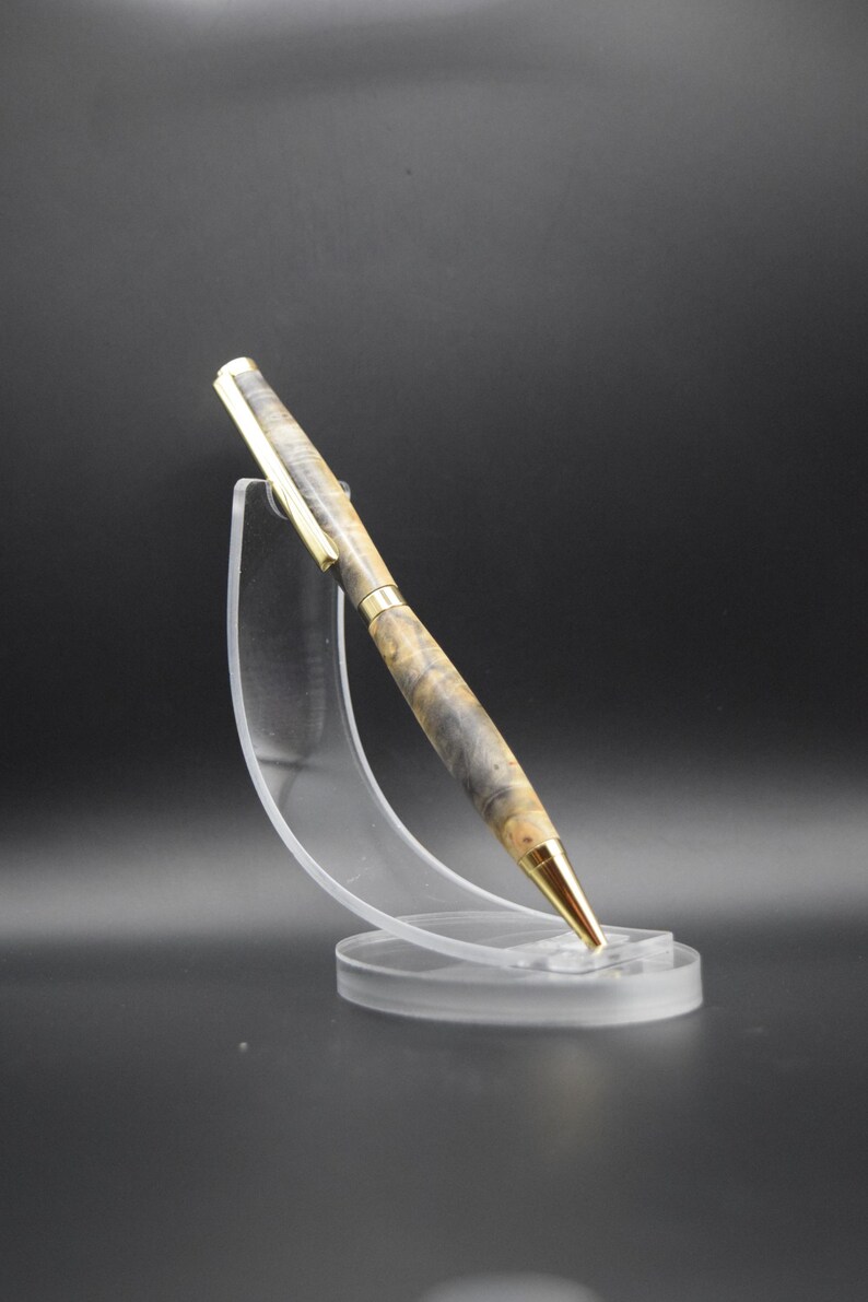 Puede incluir: Un bol&iacute;grafo artesanal con punta y detalles dorados. El cuerpo del bol&iacute;grafo presenta un patr&oacute;n &uacute;nico de vetas de madera gris y beige. Se exhibe sobre un soporte de acr&iacute;lico transparente sobre un fondo oscuro.