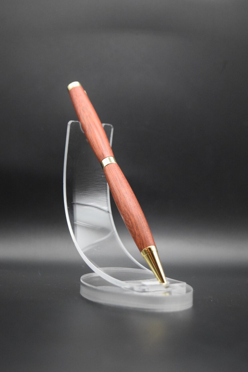 Puede incluir: Un bol&iacute;grafo de madera con acabado marr&oacute;n rojizo y detalles dorados. El bol&iacute;grafo se exhibe sobre un soporte de acr&iacute;lico transparente sobre un fondo oscuro. El dise&ntilde;o del bol&iacute;grafo presenta un cuerpo liso y pulido y una punta dorada.
