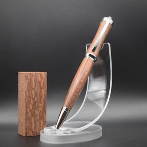 May include: A wooden pen with a silver trim and a brown wood grain pattern. The pen is displayed on a clear plastic stand.