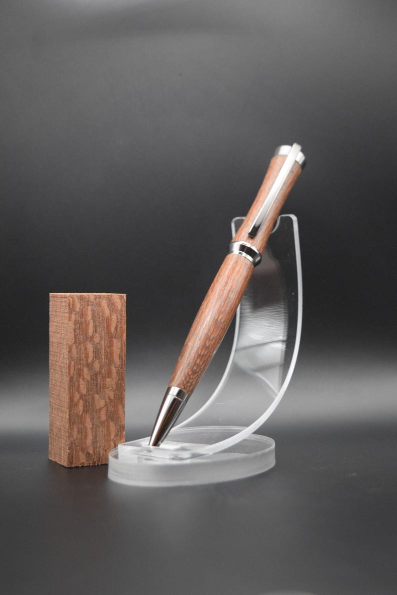 May include: A wooden pen with a silver trim and a brown wood grain pattern. The pen is displayed on a clear plastic stand.