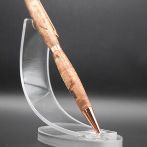 May include: A handcrafted wooden pen with copper-colored accents. The pen features a light brown, burl wood grain pattern and is displayed on a clear acrylic stand. The pen's tip, clip, and bands are copper-colored.